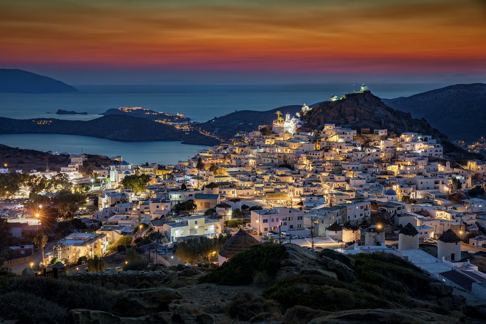 Whitewashed Greek island town illuminated at dusk on a hillside, with windmills in the foreground and a bay in the background. Ios, Greece