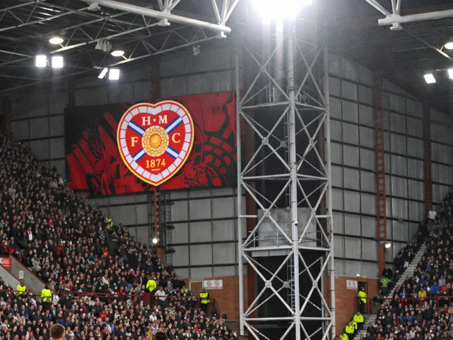 Een nachtelijke voetbalwedstrijd in een fel verlicht stadion met spelers op het veld, een groot scherm dat het Hearts F.C.-logo toont en volle tribunes.