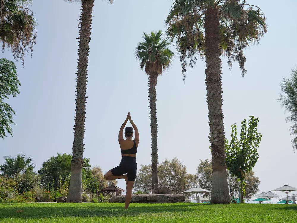 A person in Tree Pose yoga on a grassy lawn surrounded by palm trees. Esperides Beach Resort