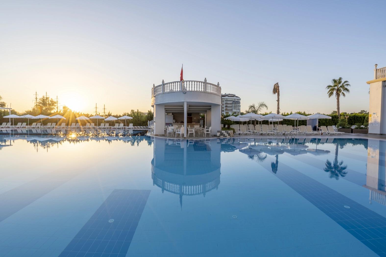 En resorts swimmingpool reflekterer en hvid poolbar, liggestole og den nedgående sol. J´adore Deluxe Hotel og Spa, Side