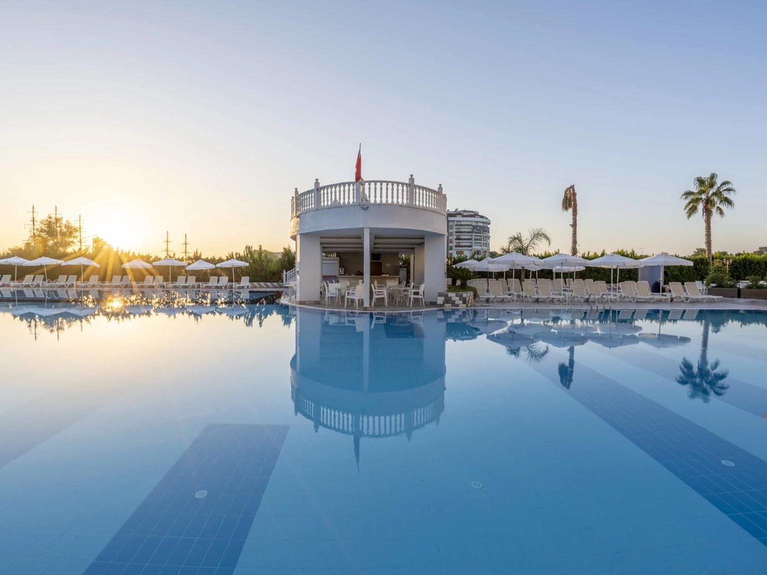 En resorts swimmingpool reflekterer en hvid poolbar, liggestole og den nedgående sol. J´adore Deluxe Hotel og Spa, Side