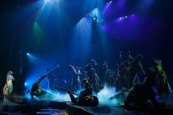 A dramatic stage scene shows a dark-robed character flying high above a crowd looking up, while a light-dressed character watches from the left. The stage is filled with blue light and smoke.