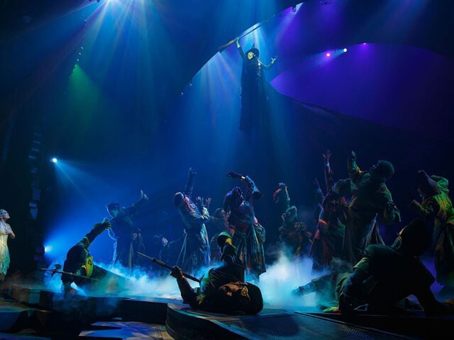 A dramatic stage scene shows a dark-robed character flying high above a crowd looking up, while a light-dressed character watches from the left. The stage is filled with blue light and smoke.