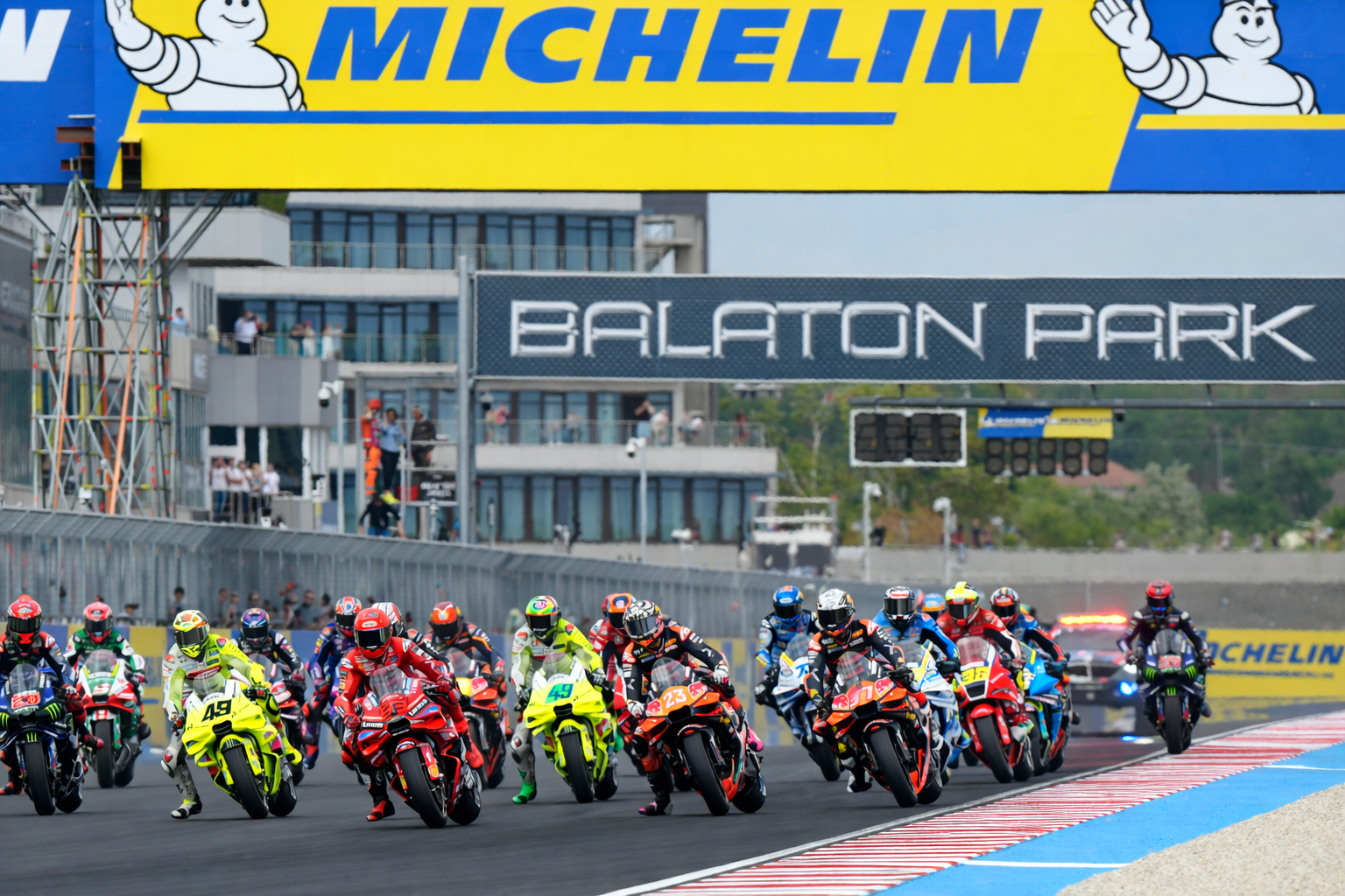 Motocykliści wyścigowi pochylają się w zakręcie na torze, nad którym wiszą banery „Michelin” i „Balaton Park”.