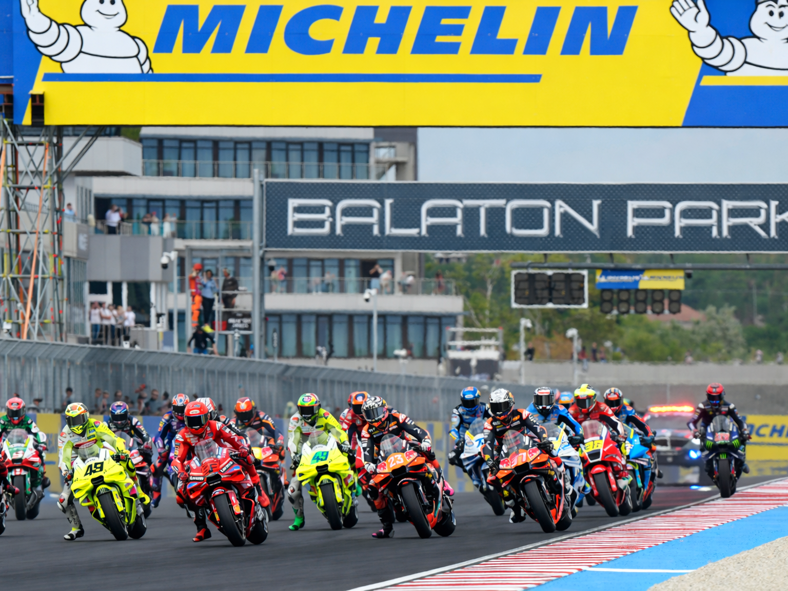 Motorcycle racers lean into a turn on a track with "Michelin" and "Balaton Park" banners overhead.