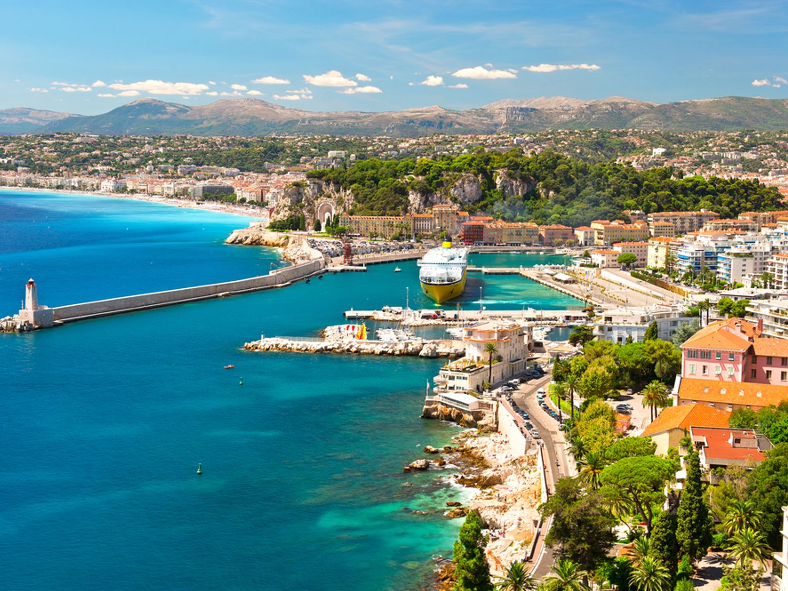 Luftaufnahme einer lebendigen Küstenhafenstadt mit einem Leuchtturm, türkisfarbenem Wasser, einem großen gelben Schiff und Bergen im Hintergrund. Nizza, Frankreich
