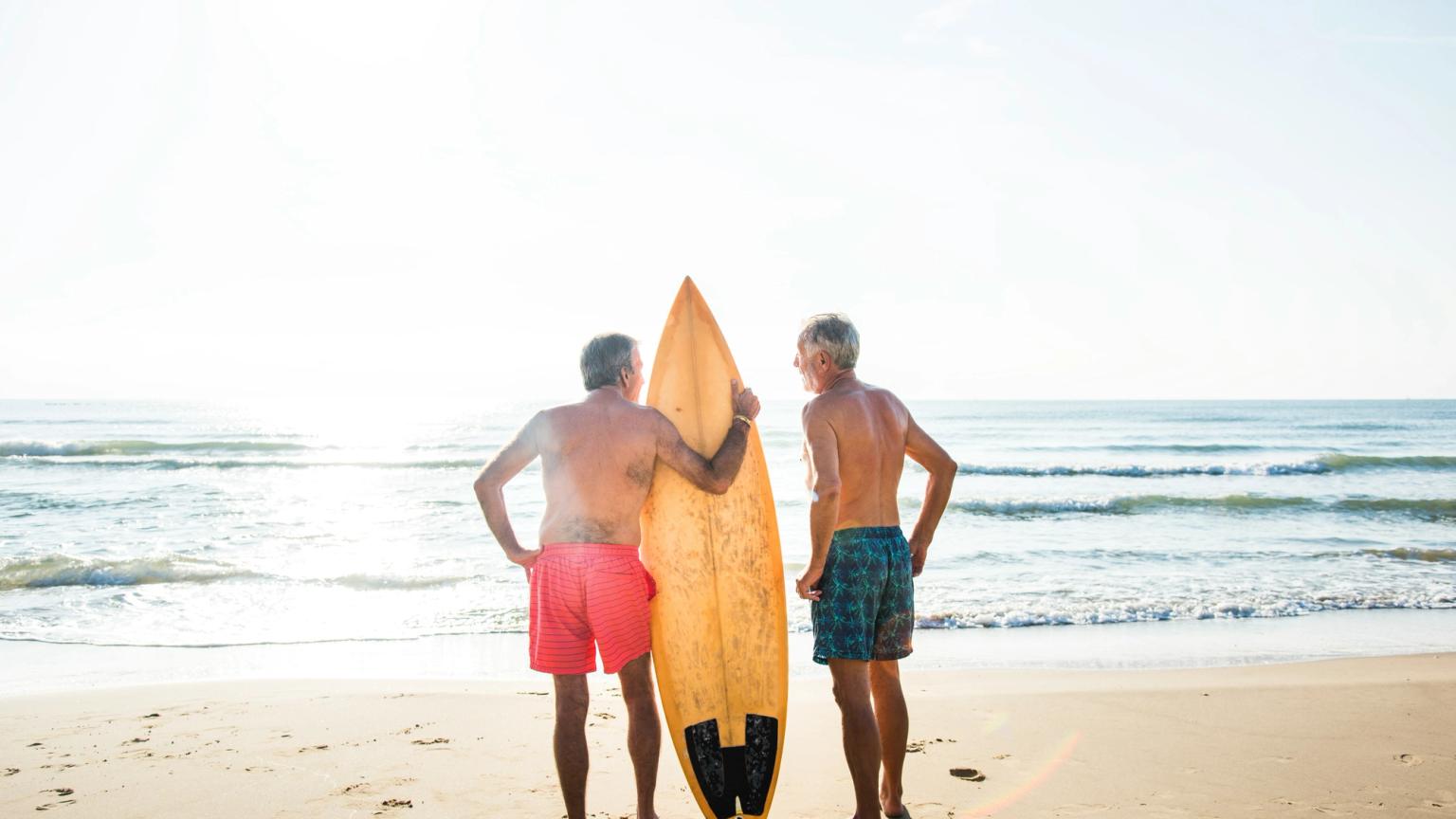 Två äldre män med en surfbräda står på en strand och tittar ut över havet.