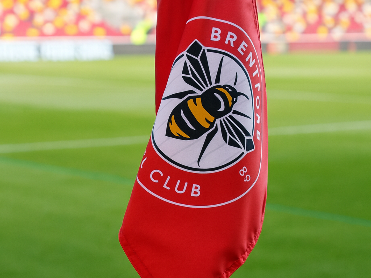 Un drapeau de coin rouge du Brentford FC avec un logo d'abeille se dresse sur un terrain de football avec des sièges de stade en arrière-plan.