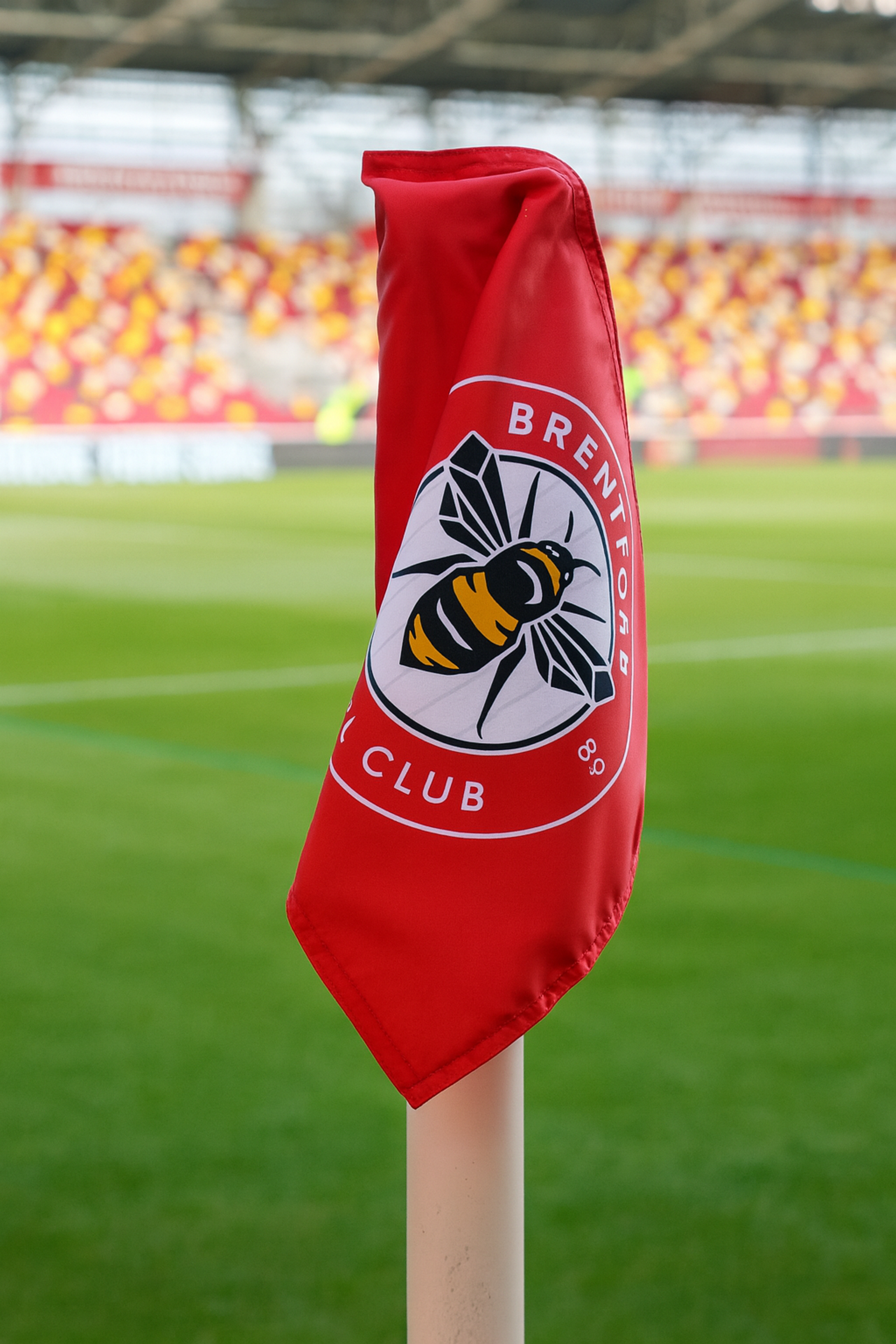 Un drapeau de coin rouge du Brentford FC avec un logo d'abeille se dresse sur un terrain de football avec des sièges de stade en arrière-plan.