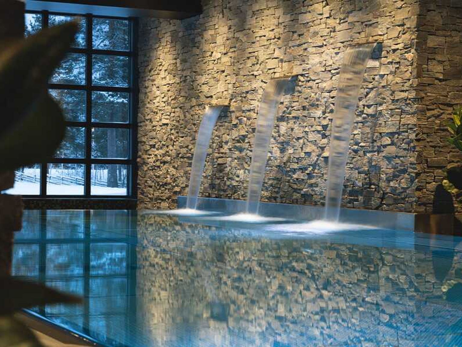 Indendørs pool med sten vandfaldsvæg, stort vindue der viser et sneklædt landskab og stemningsbelysning.