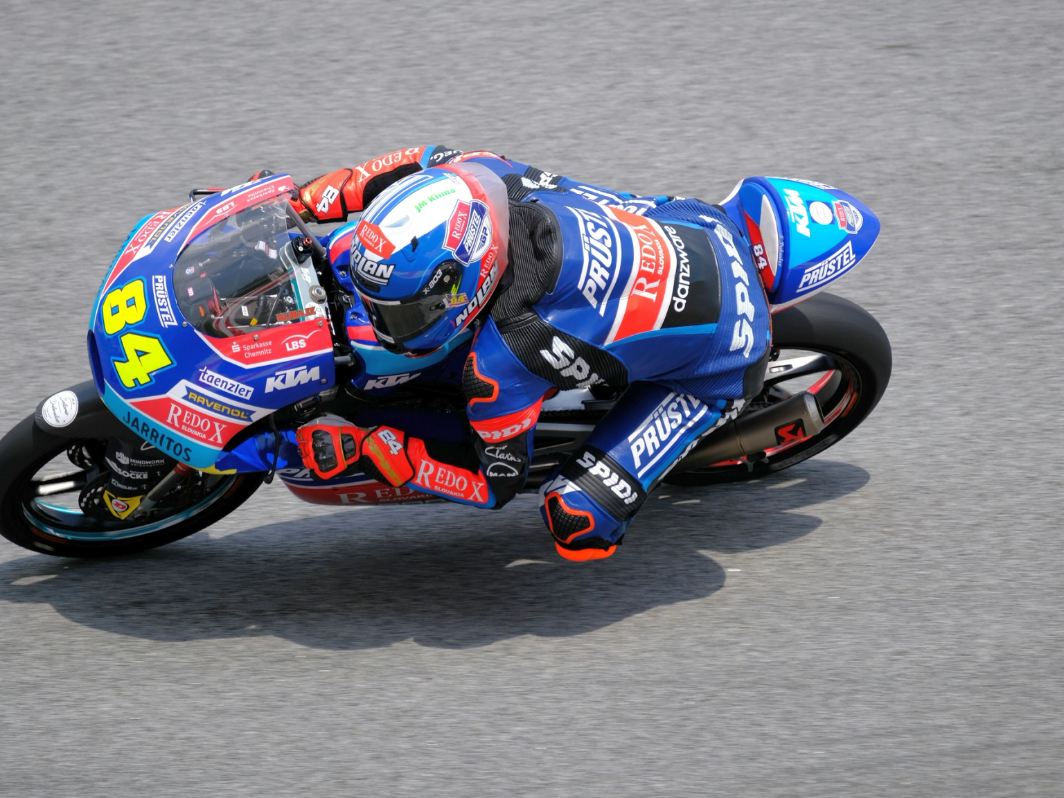 Motorcycle racer number 84 in a blue suit leans sharply into a turn on a blue and white bike.