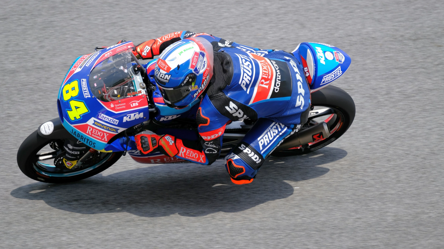 Motorcycle racer number 84 in a blue suit leans sharply into a turn on a blue and white bike.