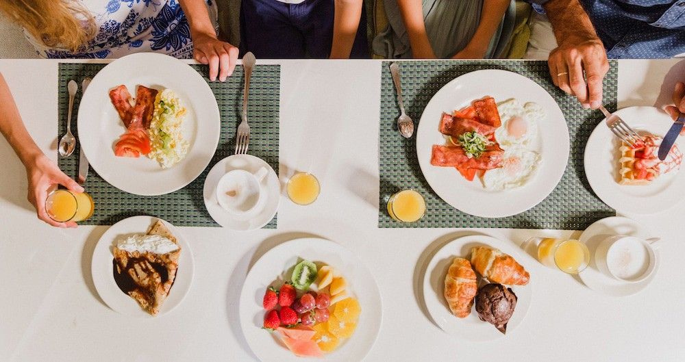 Draufsicht auf einen Frühstückstisch mit Tellern mit Eiern, Speck, Obst, Gebäck, Waffeln und Saft, bei dem die Hände der Personen sichtbar sind. Aparthotel Club del Sol Resort & SPA