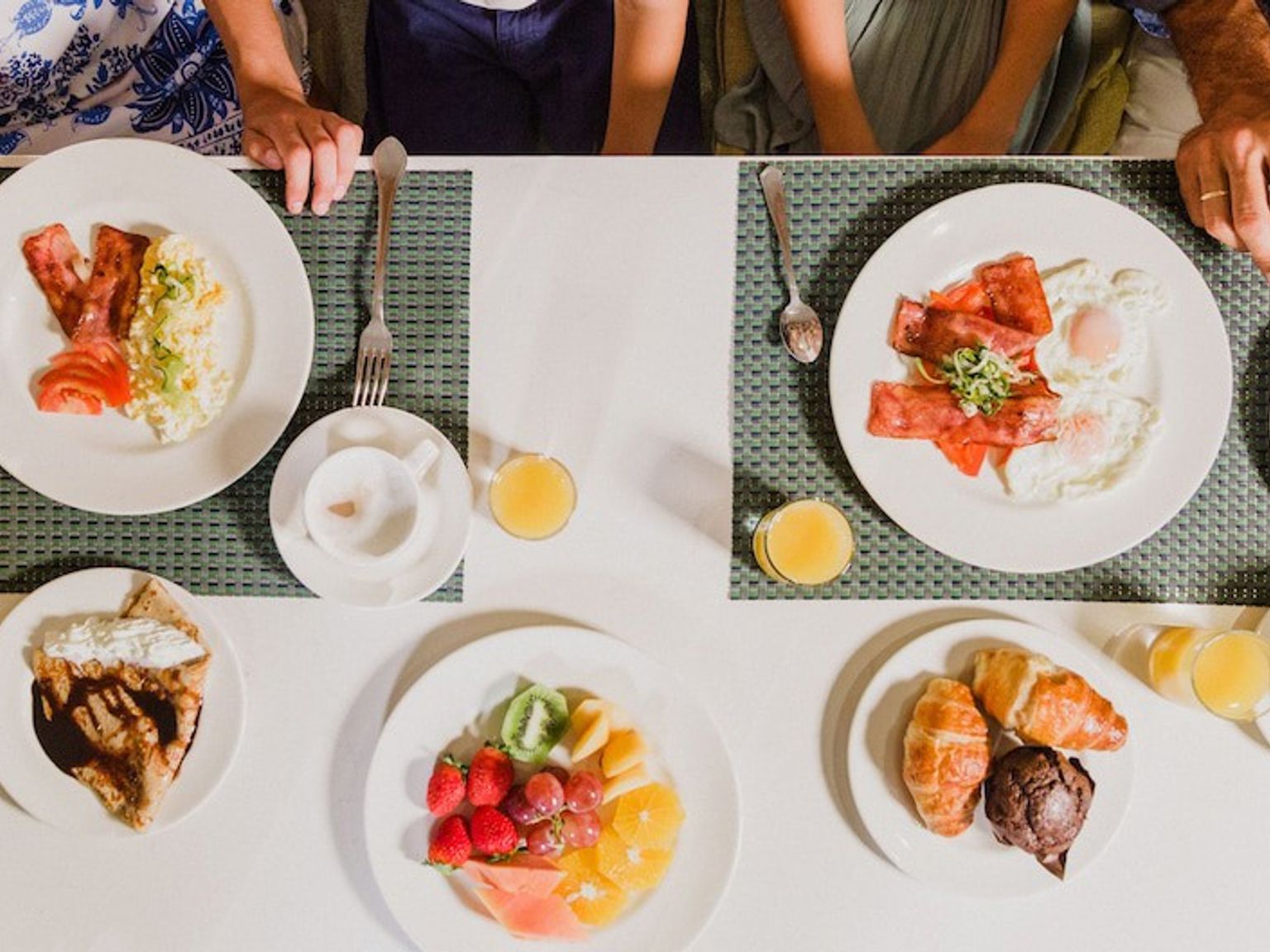 Draufsicht auf einen Frühstückstisch mit Tellern mit Eiern, Speck, Obst, Gebäck, Waffeln und Saft, bei dem die Hände der Personen sichtbar sind. Aparthotel Club del Sol Resort & SPA