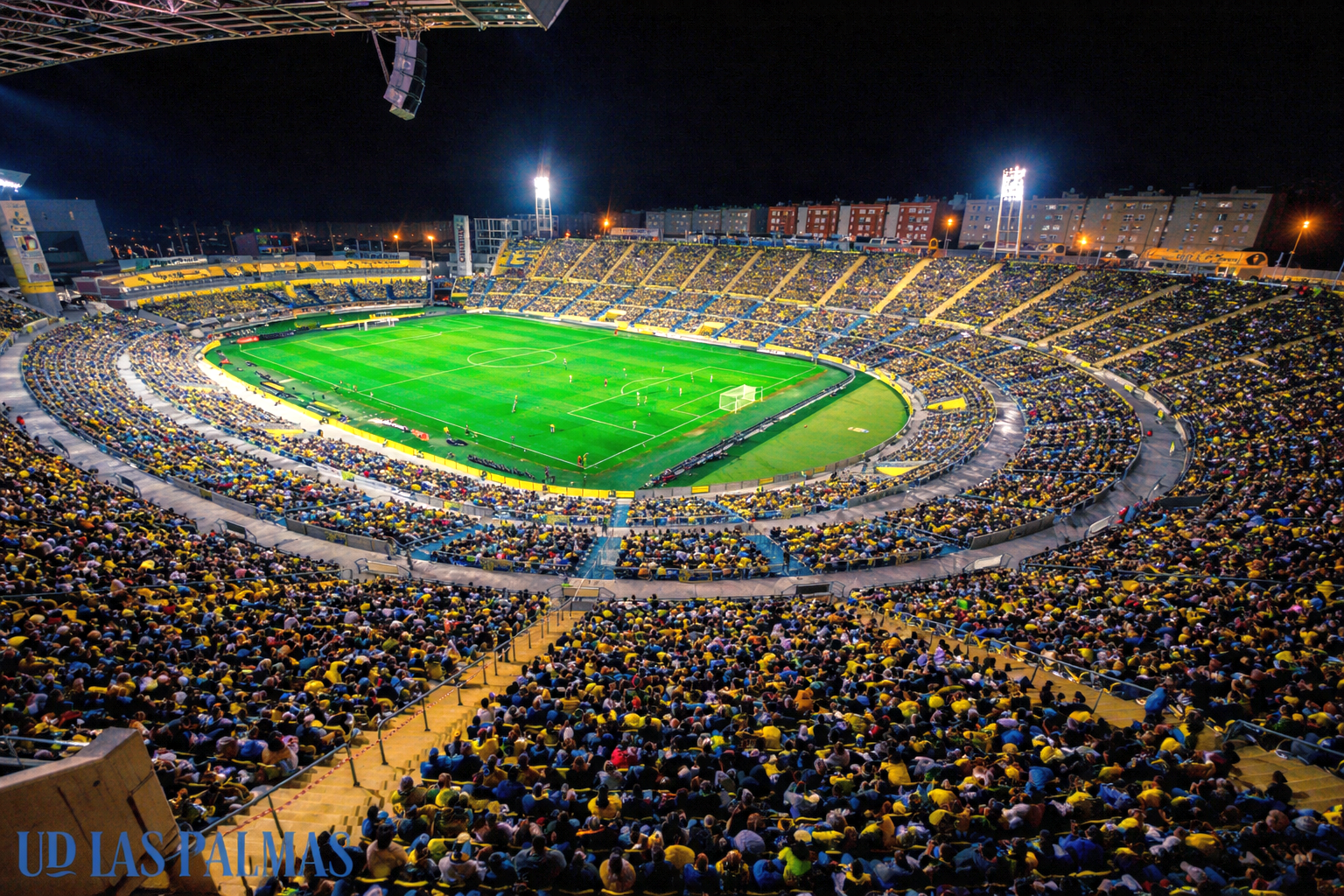 Un estadio de fútbol lleno por la noche con un campo verde brillantemente iluminado y aficionados en amarillo y azul.