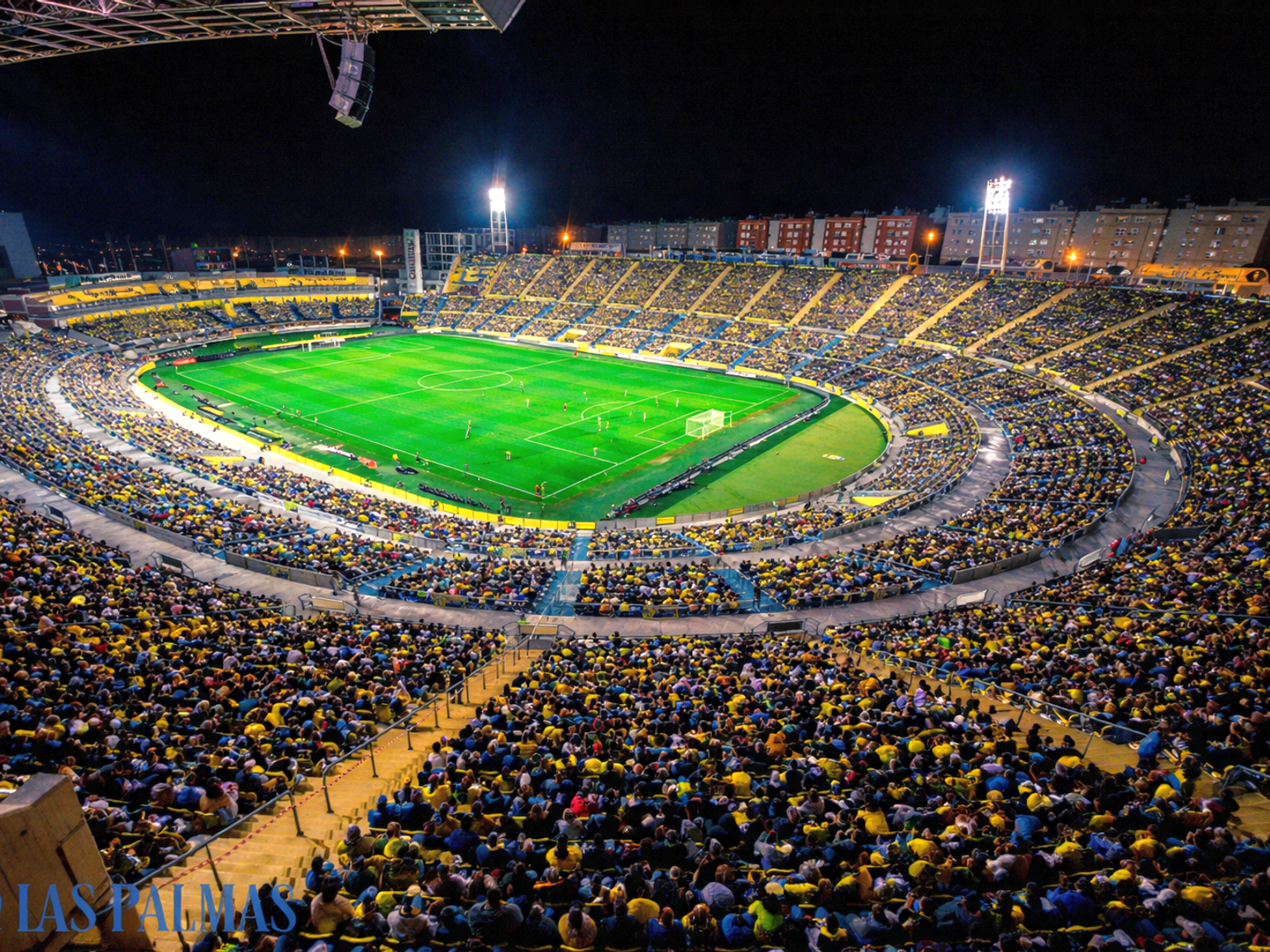 Un estadio de fútbol lleno por la noche con un campo verde brillantemente iluminado y aficionados en amarillo y azul.
