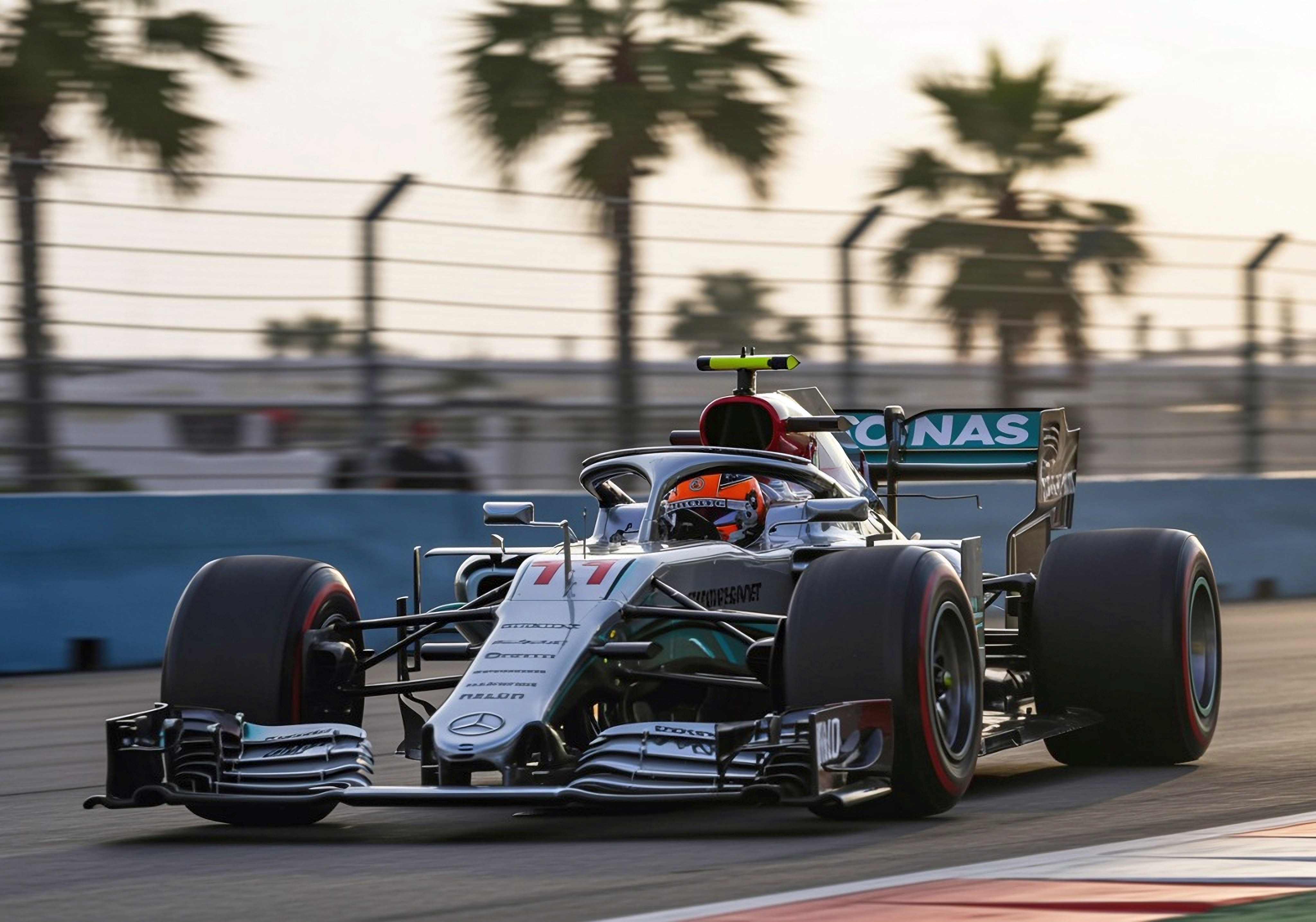 A Mercedes F1 car, number 77, races on a track with palm trees in the blurred background.