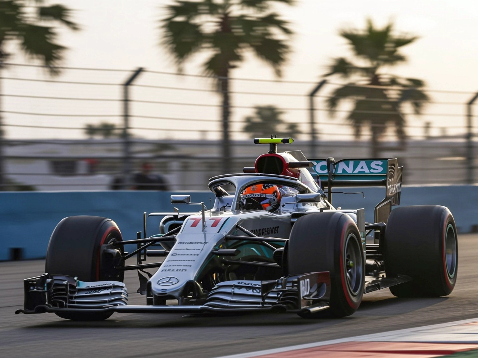 A Mercedes F1 car, number 77, races on a track with palm trees in the blurred background.
