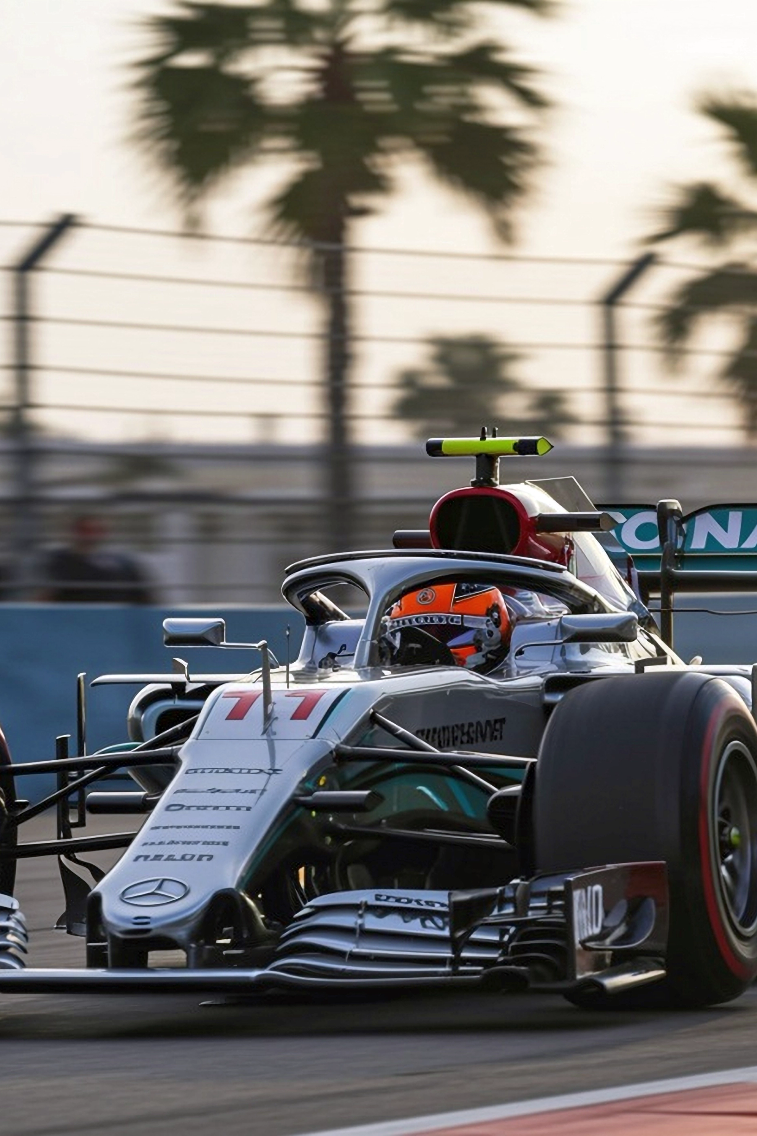 A Mercedes F1 car, number 77, races on a track with palm trees in the blurred background.