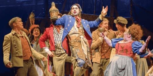 An energetic stage performance featuring actors in period costumes, with a man in a blue jacket holding a bottle high.