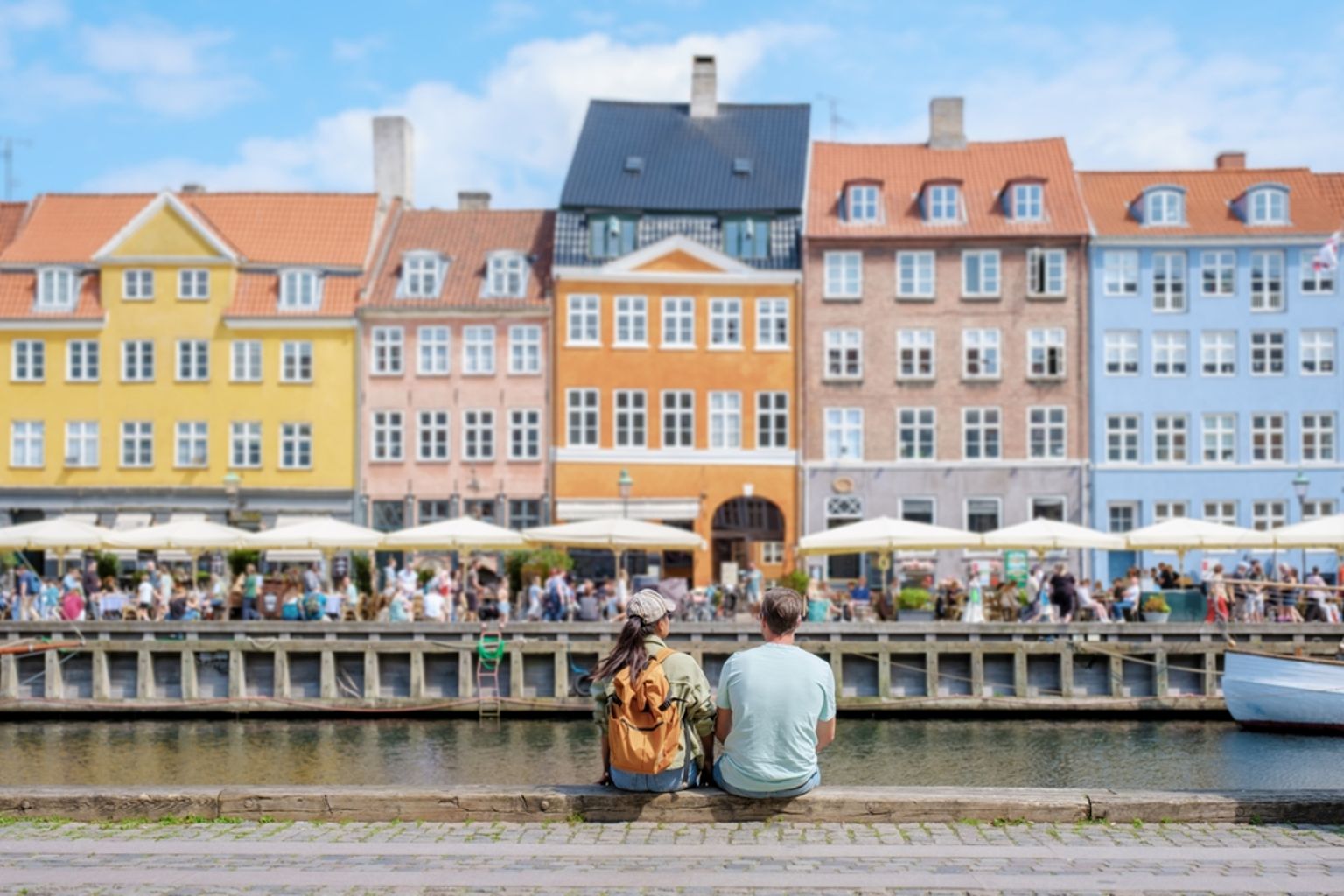 a man and a woman are sitting next to a river in front of a row of buildings.