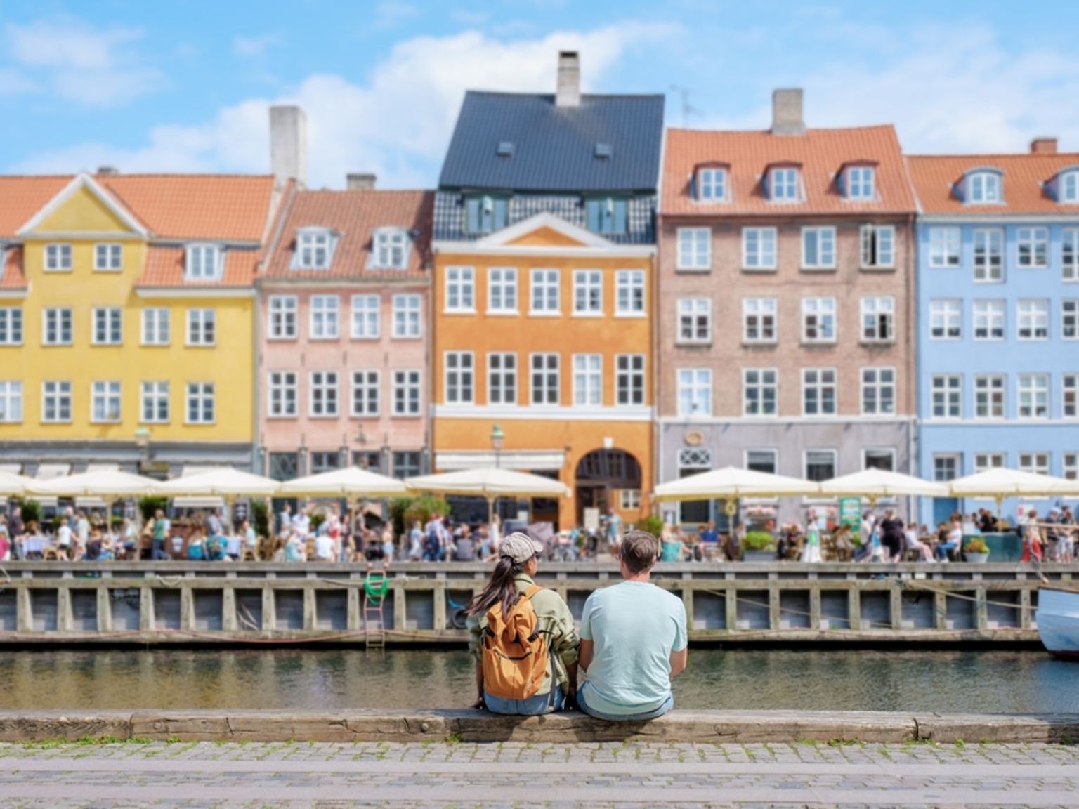 a man and a woman are sitting next to a river in front of a row of buildings.