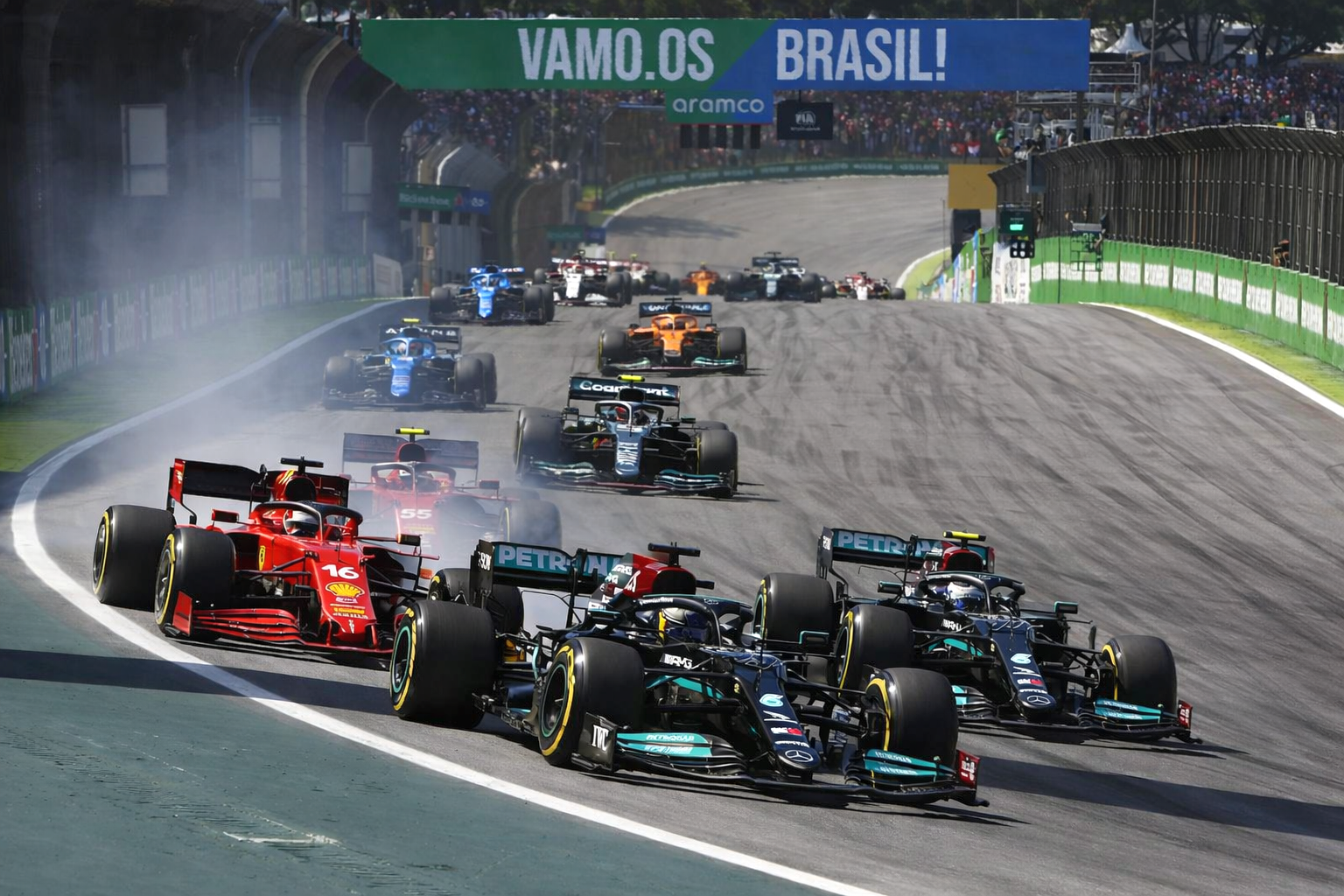 Formula 1 cars race on a track, with a red Ferrari and a black Mercedes leading the pack, kicking up tire smoke against a backdrop of spectators.