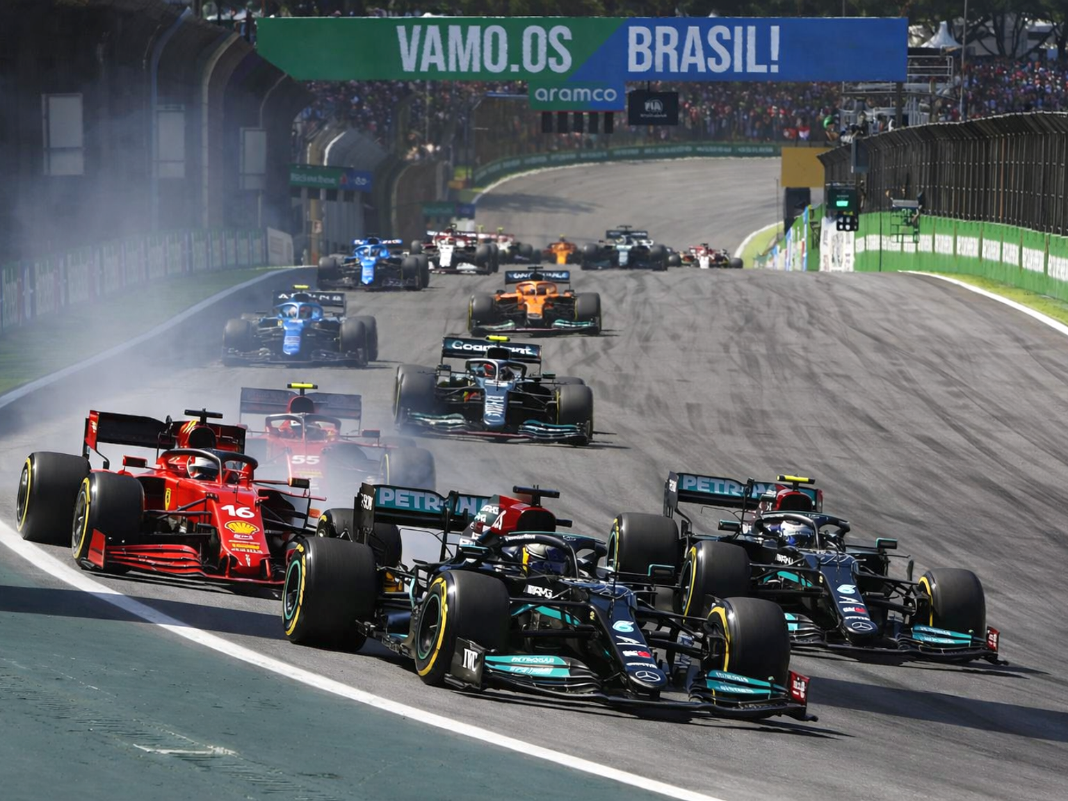 Formula 1 cars race on a track, with a red Ferrari and a black Mercedes leading the pack, kicking up tire smoke against a backdrop of spectators.