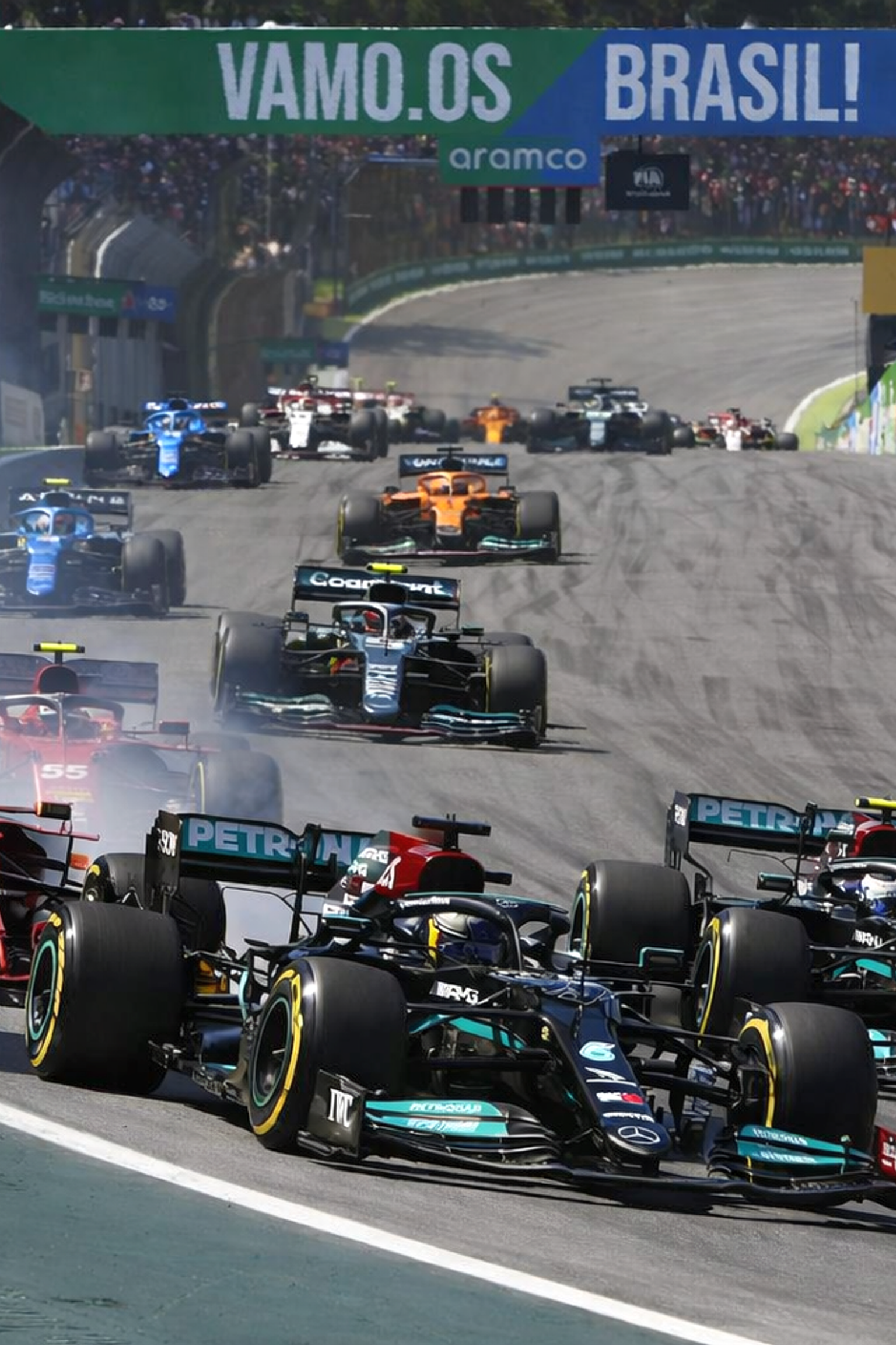 Formula 1 cars race on a track, with a red Ferrari and a black Mercedes leading the pack, kicking up tire smoke against a backdrop of spectators.