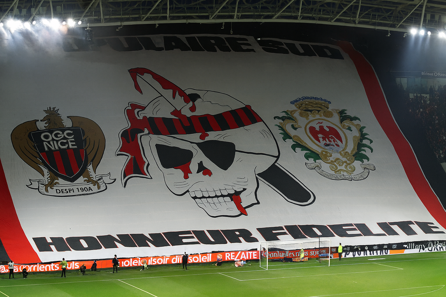 Grande tifo de estádio com o brasão do OGC Nice, um crânio sangrento com uma faca, o emblema da cidade de Nice e o texto "POPULAIRE SUD" e "HONNEUR FIDELITE."
