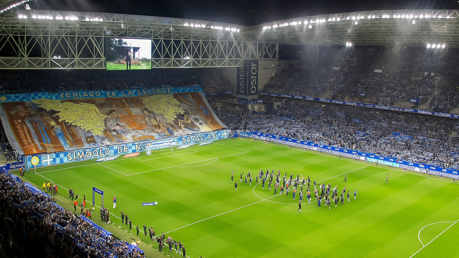 Een verlicht voetbalstadion 's nachts, gevuld met fans, heeft een enorm blauw en goud tifo-banner met Spaanse tekst, en spelers verzameld op het groene veld.