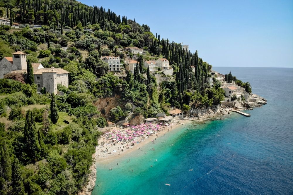 A vibrant beach with pink umbrellas and swimmers in a turquoise bay, overlooked by a lush hillside village. Dubrovnik, Croatia