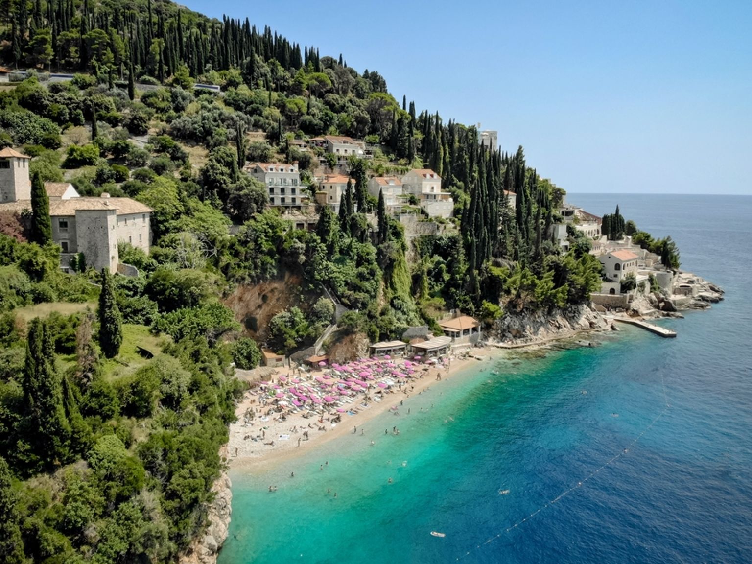 En livlig strand med lyserøde parasoller og svømmere i en turkis bugt, med udsigt til en frodig landsby på en bjergskråning. Dubrovnik, Kroatien