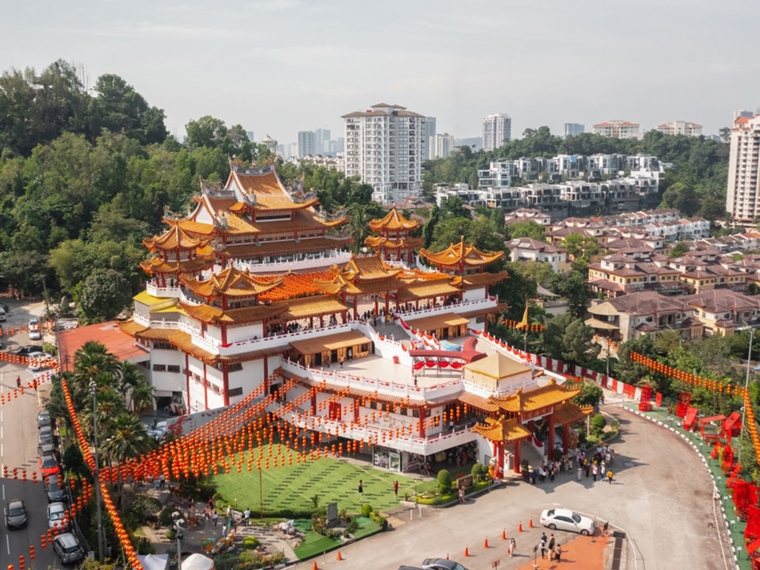 Et stort, udsmykket tempel med orange tag i flere niveauer og adskillige røde lanterner, placeret mod en grøn bakke og bybaggrund. Kuala Lumpur