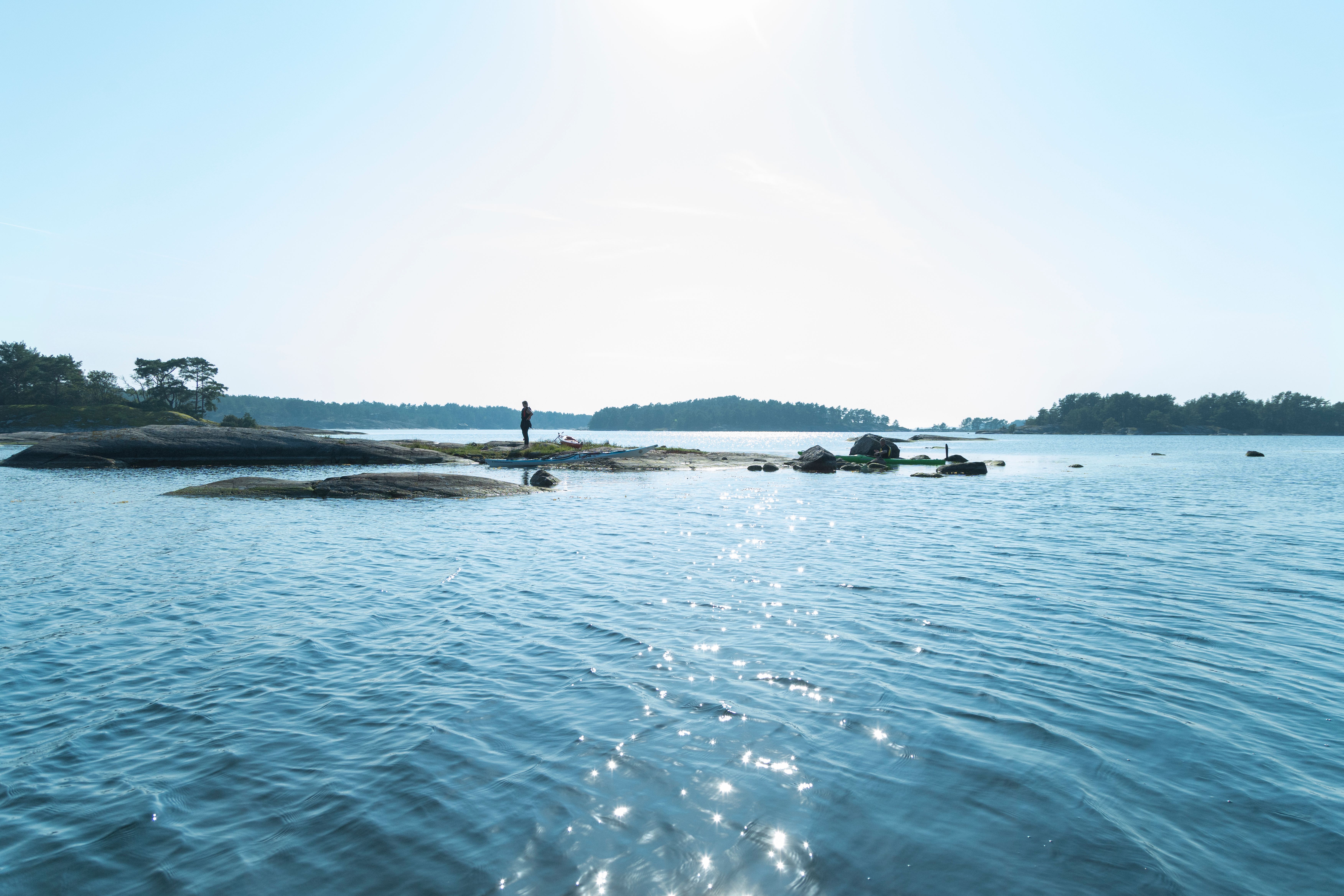 En person står på en lille, klippefyldt ø i en rolig, solplettet bugt, med andre træklædte øer i det fjerne.