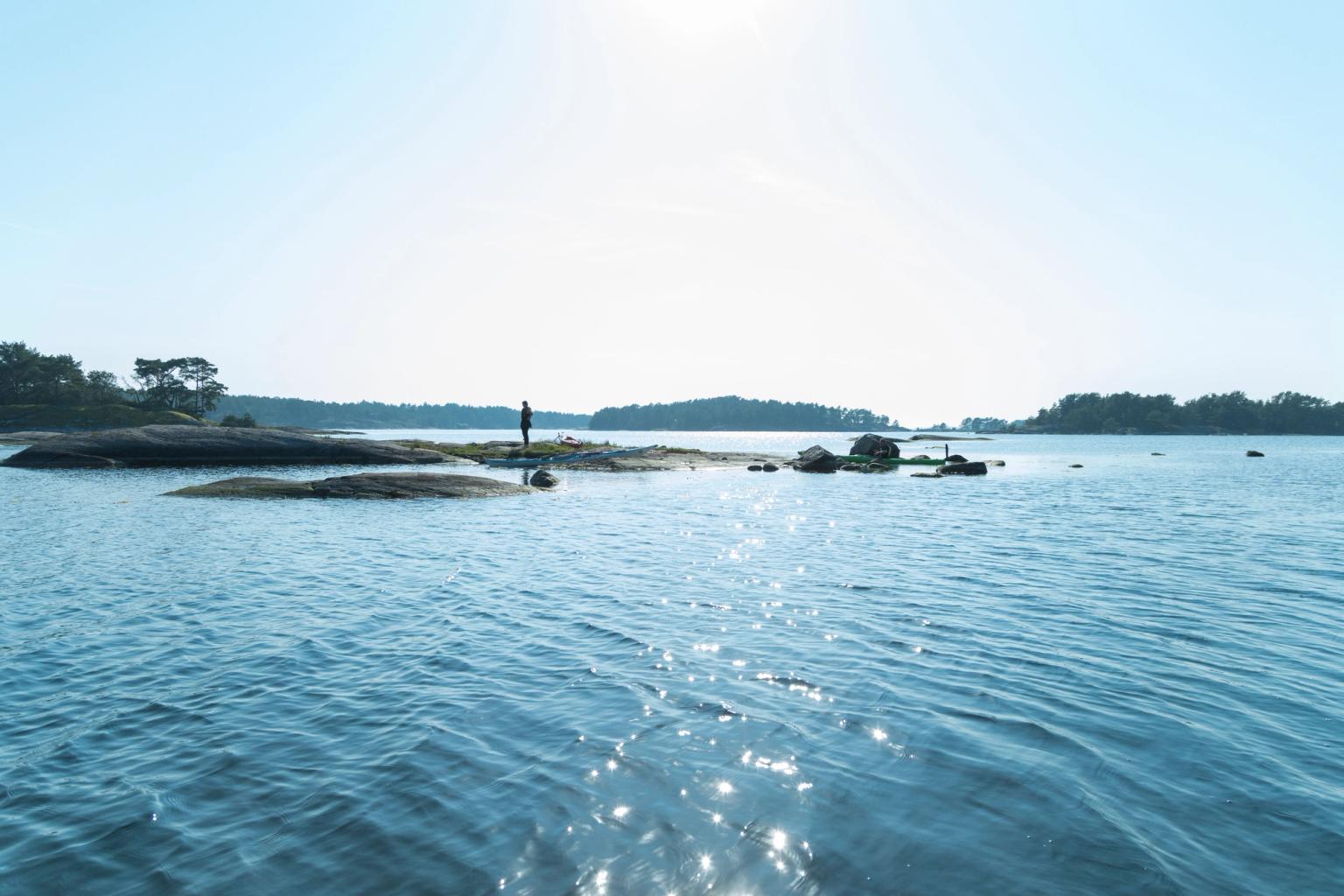 En person står på en lille, klippefyldt ø i en rolig, solplettet bugt, med andre træklædte øer i det fjerne.