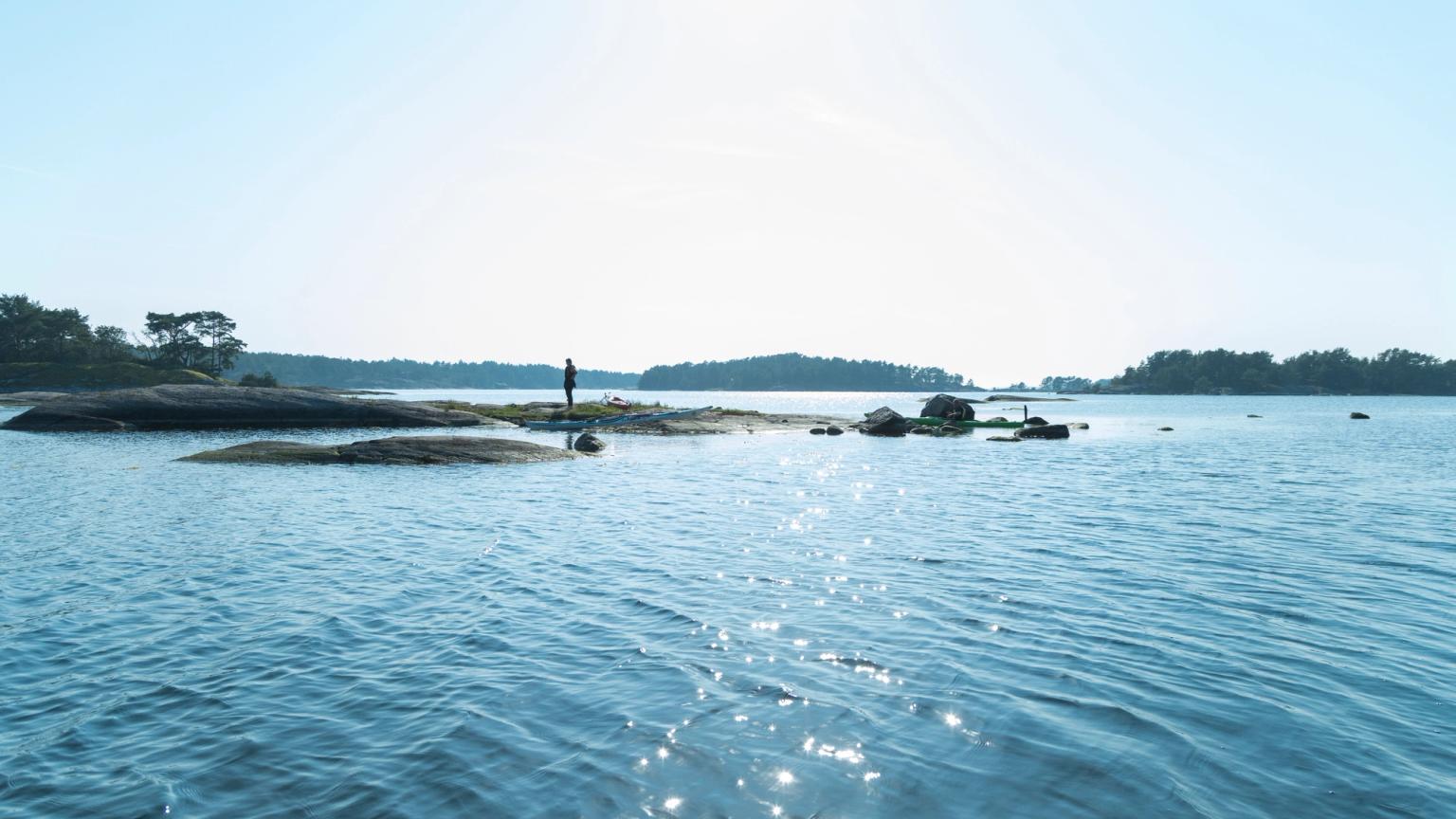 En person står på en lille, klippefyldt ø i en rolig, solplettet bugt, med andre træklædte øer i det fjerne.