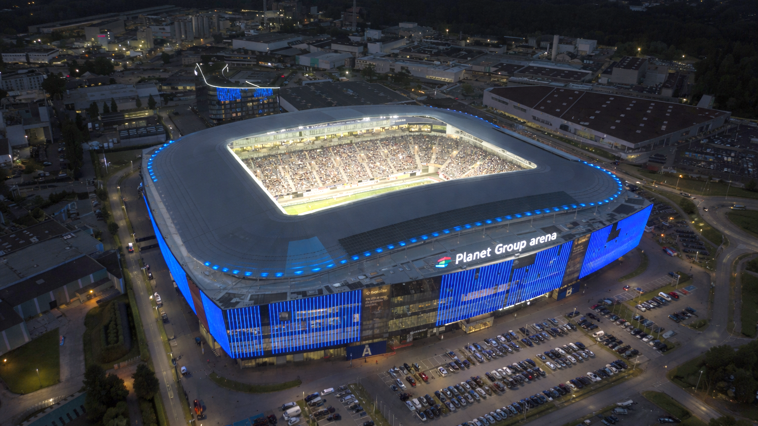 Aerial view of Planet Group Arena at night, brightly lit with blue exterior lights and an interior full of spectators, surrounded by a parking lot and industrial buildings.