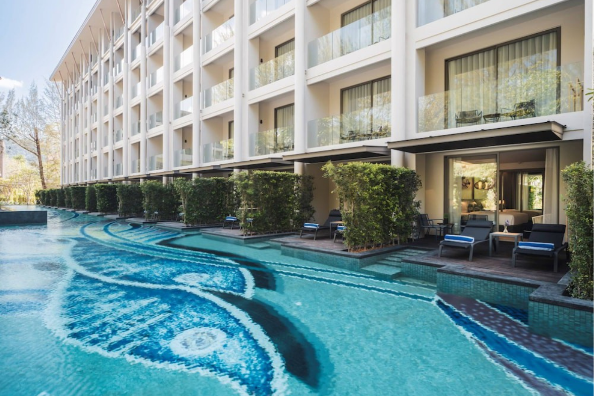 En modern resortbyggnad med balkonger som vetter mot en lång, slingrande pool med ett blått mosaikmönster. Rummen på bottenvåningen har uteplatser med solstolar och direkt tillgång till poolen.