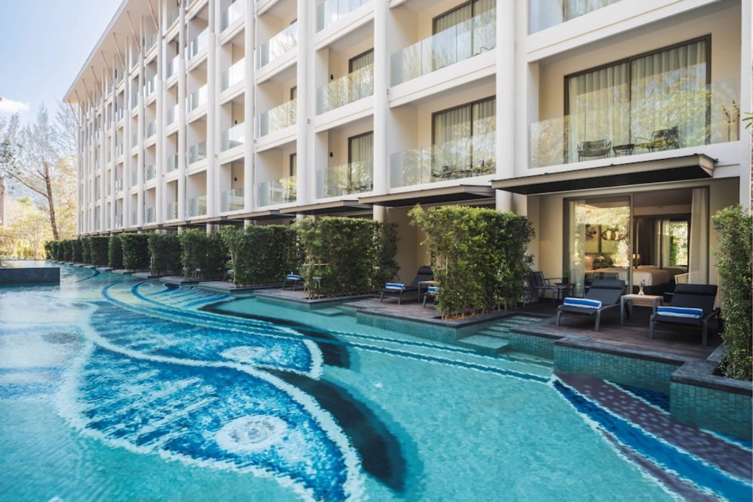 En modern resortbyggnad med balkonger som vetter mot en lång, slingrande pool med ett blått mosaikmönster. Rummen på bottenvåningen har uteplatser med solstolar och direkt tillgång till poolen.