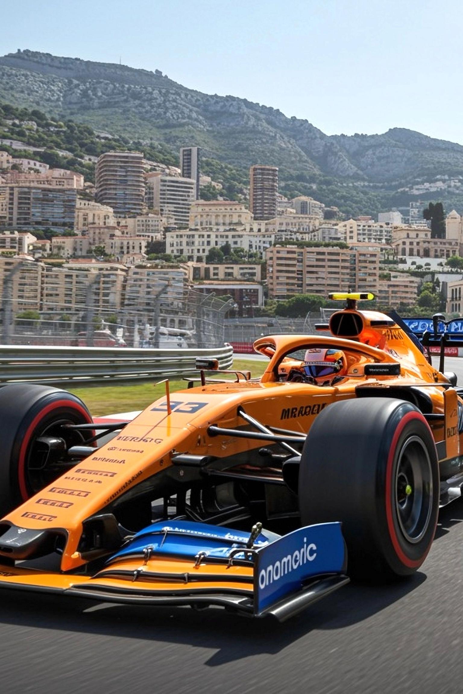 An orange and blue McLaren Formula 1 car drives on a track with a city built into a mountainside in the background.