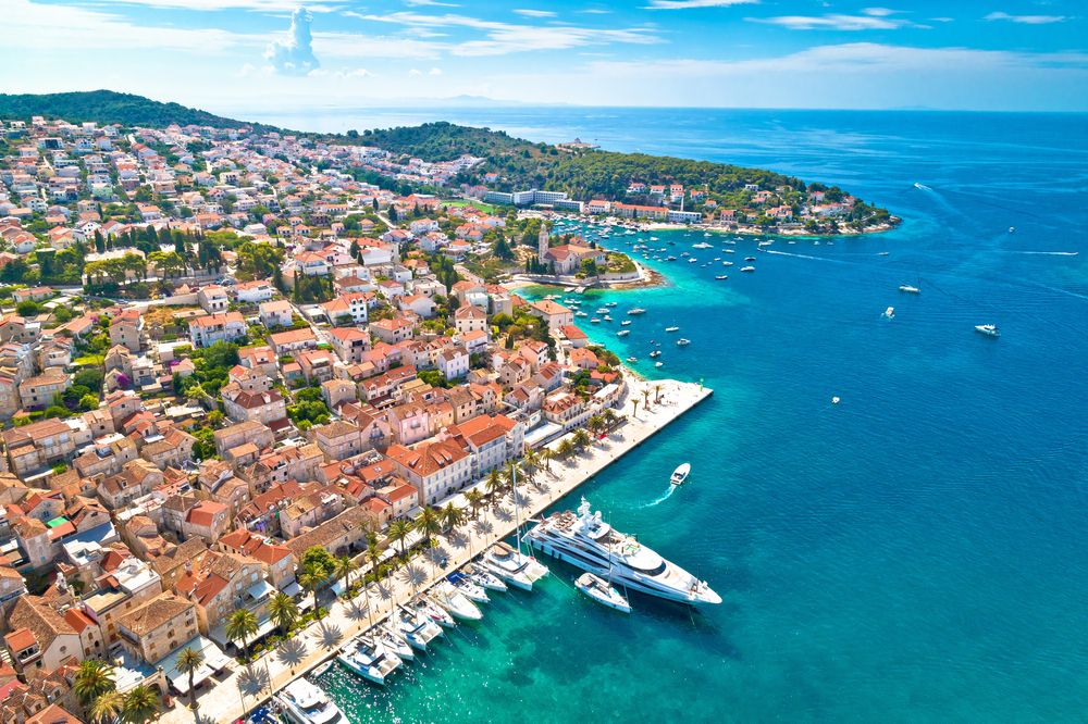 Luftfoto af en kystby med røde tegltage, en travl havn med både og klart blåt vand. Hvar, Kroatien