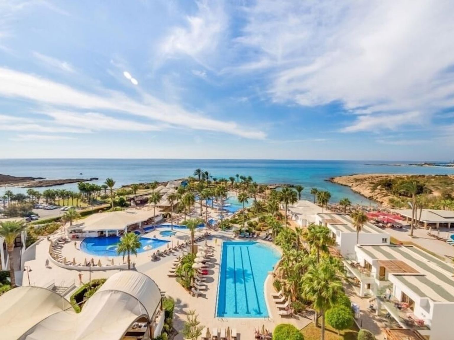 Luftfoto av et kystresort med flere bassenger, palmer og bygninger ved siden av det blå havet.
