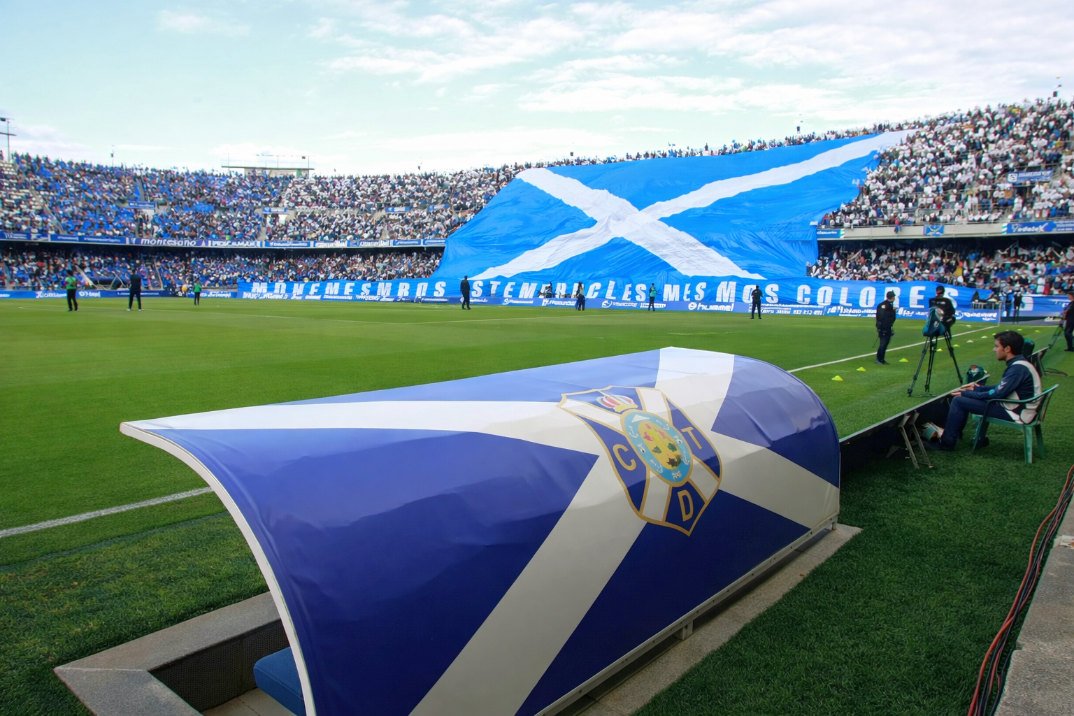Een voetbalstadion met een CD Tenerife dug-out en een grote menigte die een gigantische blauw-witte vlag met een witte saltire vasthoudt.