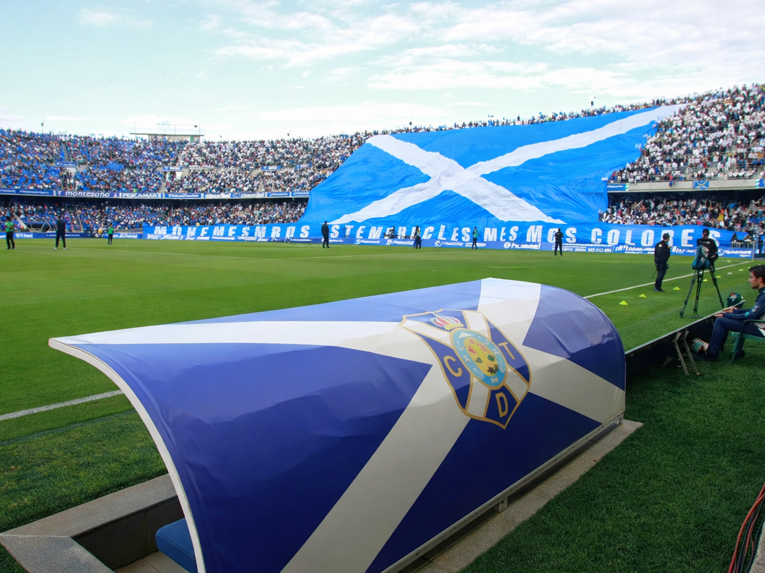 Een voetbalstadion met een CD Tenerife dug-out en een grote menigte die een gigantische blauw-witte vlag met een witte saltire vasthoudt.