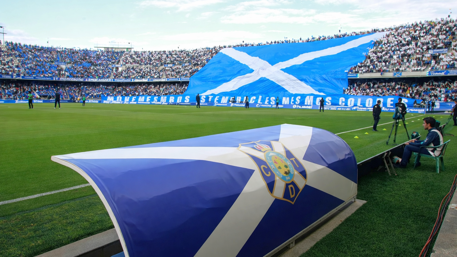 Een voetbalstadion met een CD Tenerife dug-out en een grote menigte die een gigantische blauw-witte vlag met een witte saltire vasthoudt.