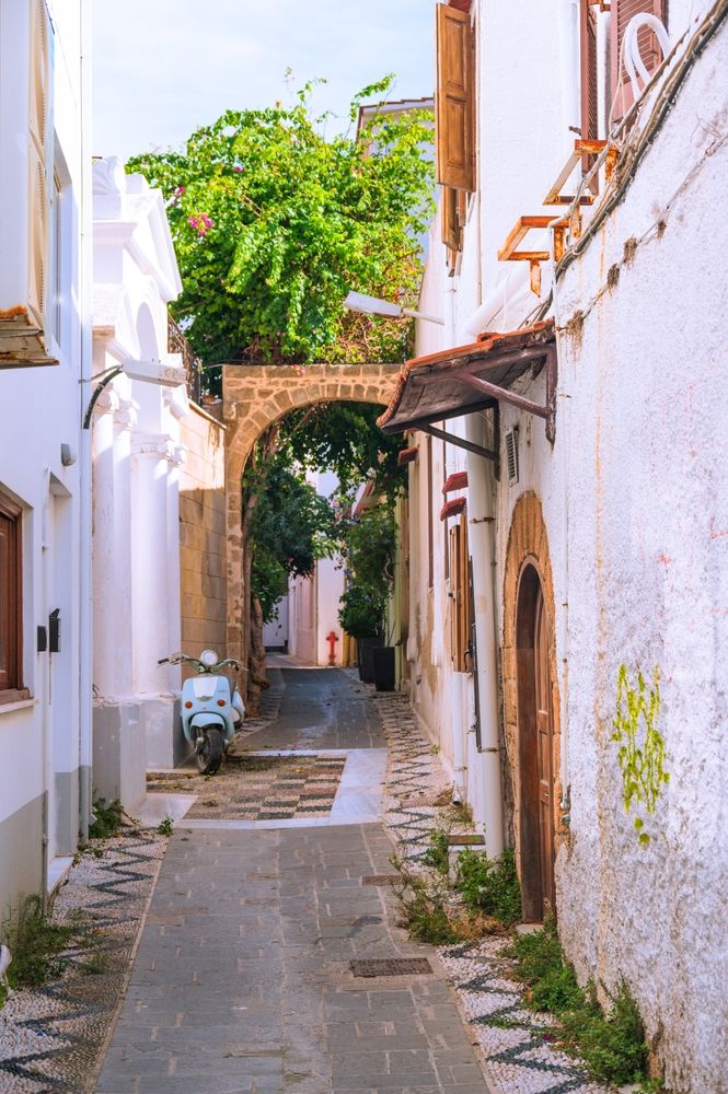 A narrow, paved alleyway lined with white buildings and lush greenery, featuring a light blue scooter on the left and a stone archway in the distance.