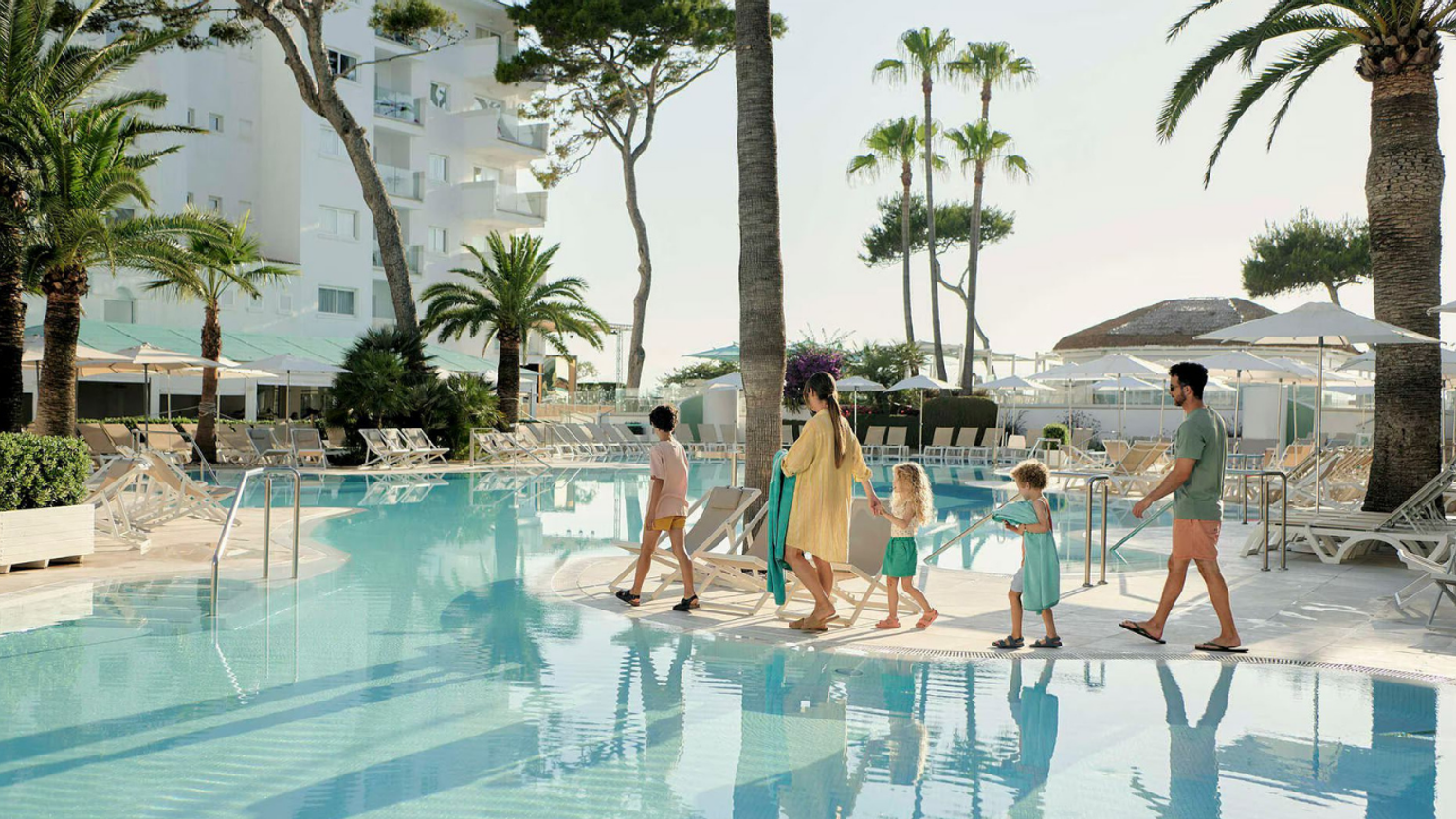 En familj på fem går längs kanten av en stor pool på en solig resort.