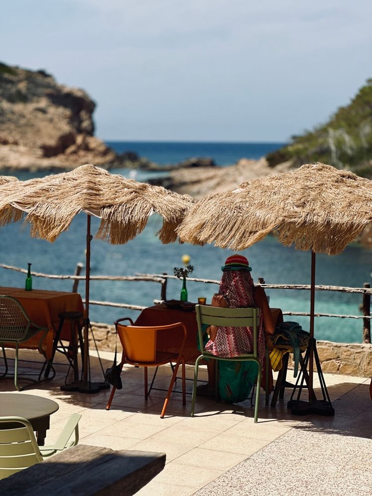 En person sitter vid ett utomhuscafébord under ett stråparasoll med utsikt över ett blått hav och en klippig kust. Ibiza, Spanien
