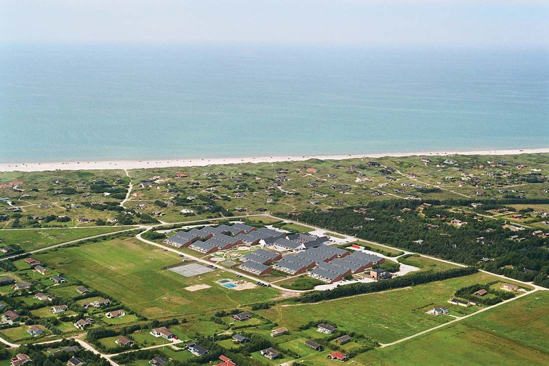 Luftfoto av et stort resortkompleks nær en sandstrand og havet, omgitt av spredte hus og grønne jorder. Landal Grønhøj Strand